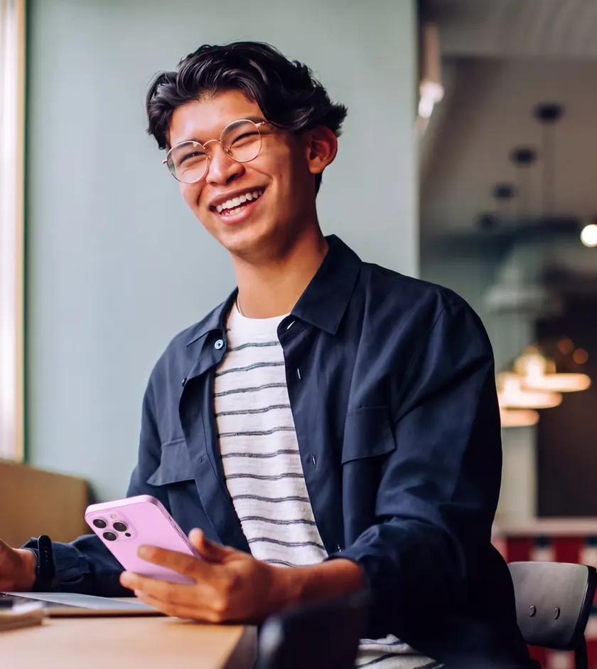 A student laughing at a café table, holding a pink phone