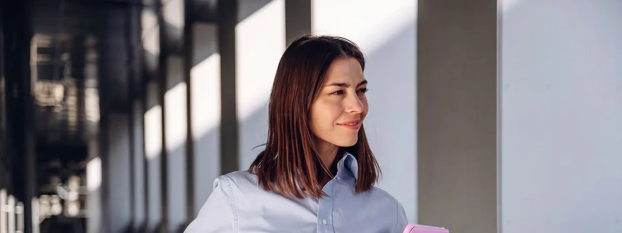 A professional in a bright corridor, holding a notebook.