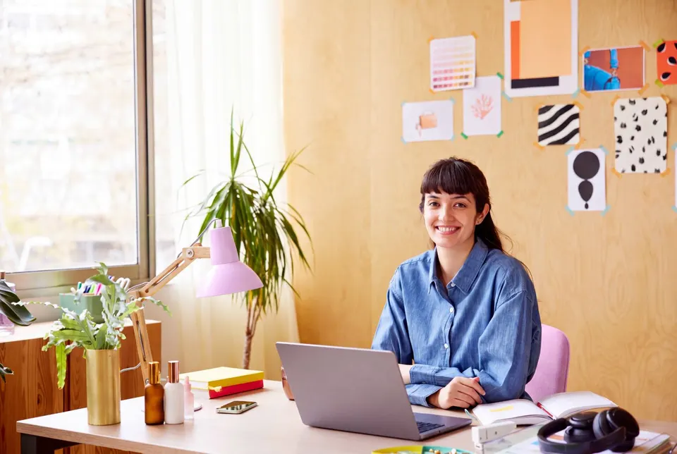 A student smiling at her desk in a bright studio, laptop open, notebook and pinned work beside her.