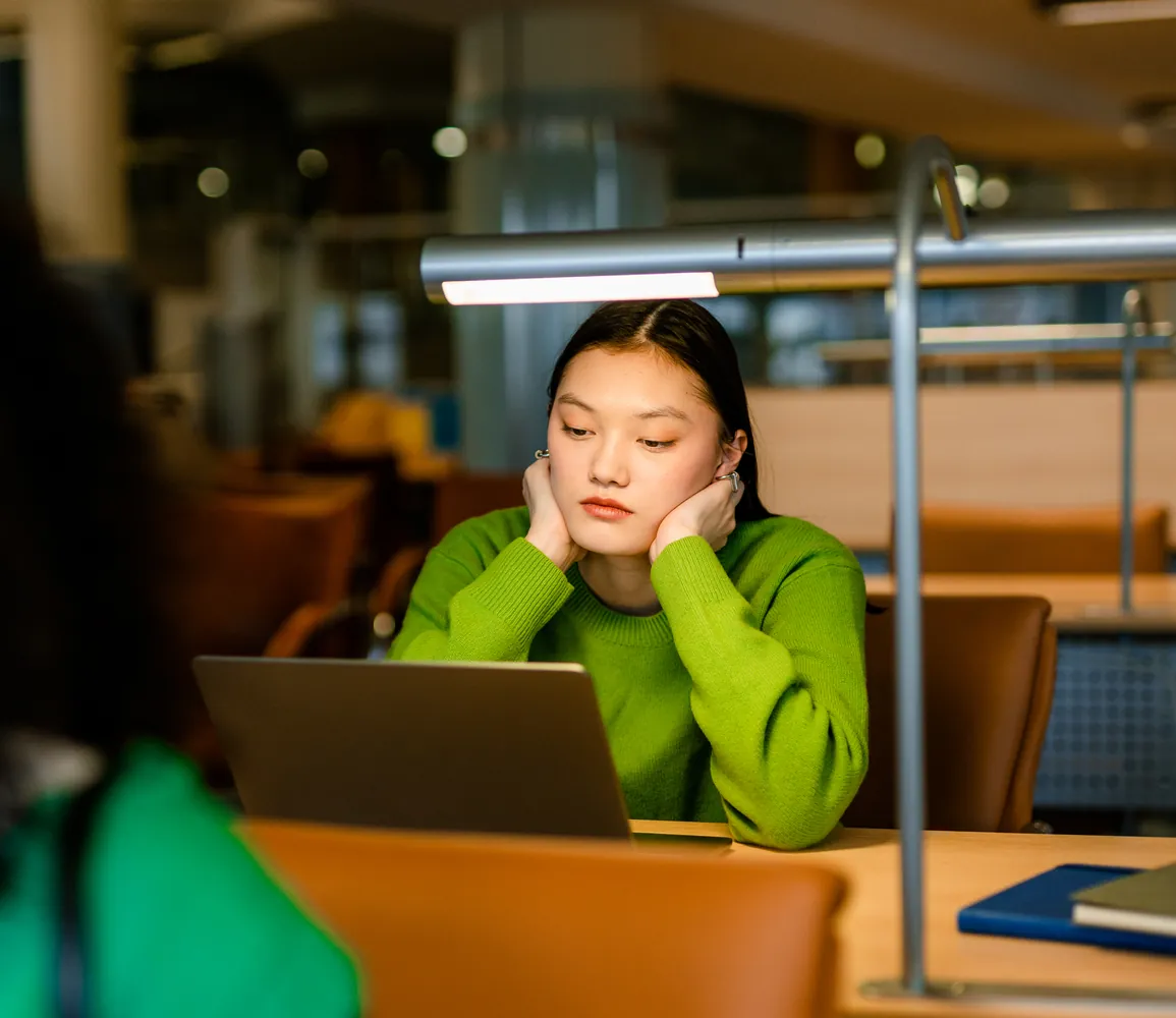 A student in a green jumper thinking over her laptop in a library