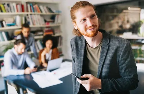 A smiling man with a phone stands in front of colleagues working at a table.